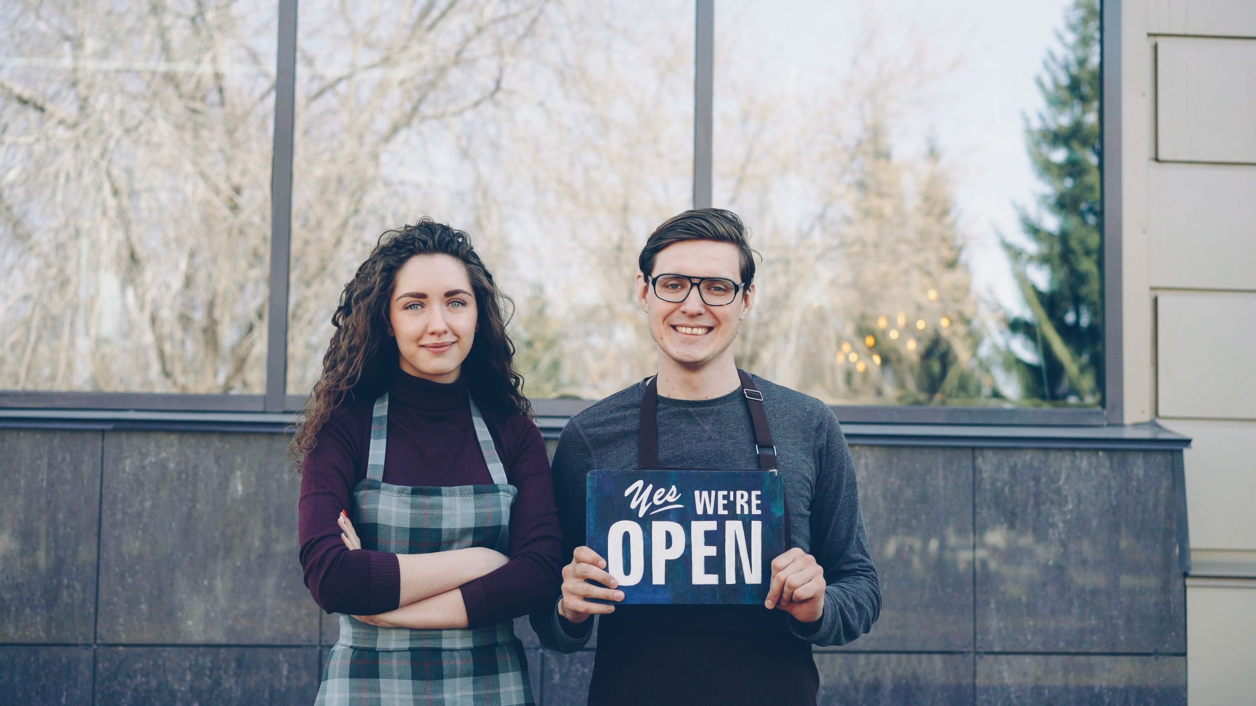 Two local business owners standing outside their shop holding a 'Yes We're Open' sign, welcoming customers
